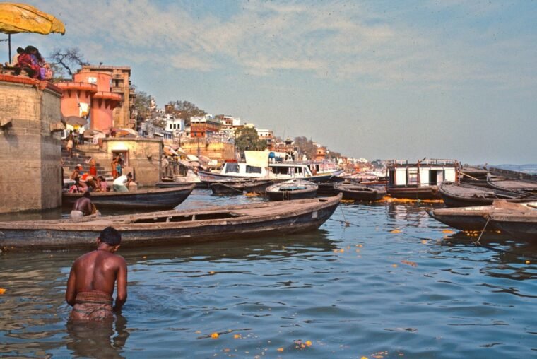 Man bathing in ganges river with boats and buildings.